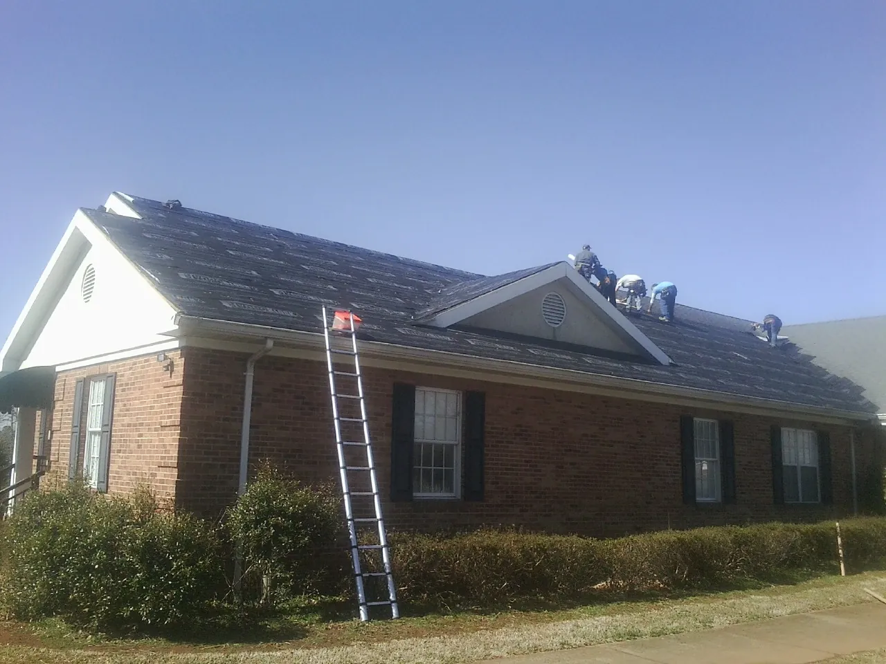 Crew tearing shingles off a brick ranch roof with a ladder set — Emergency Roof Repair work by our crew in Lancing